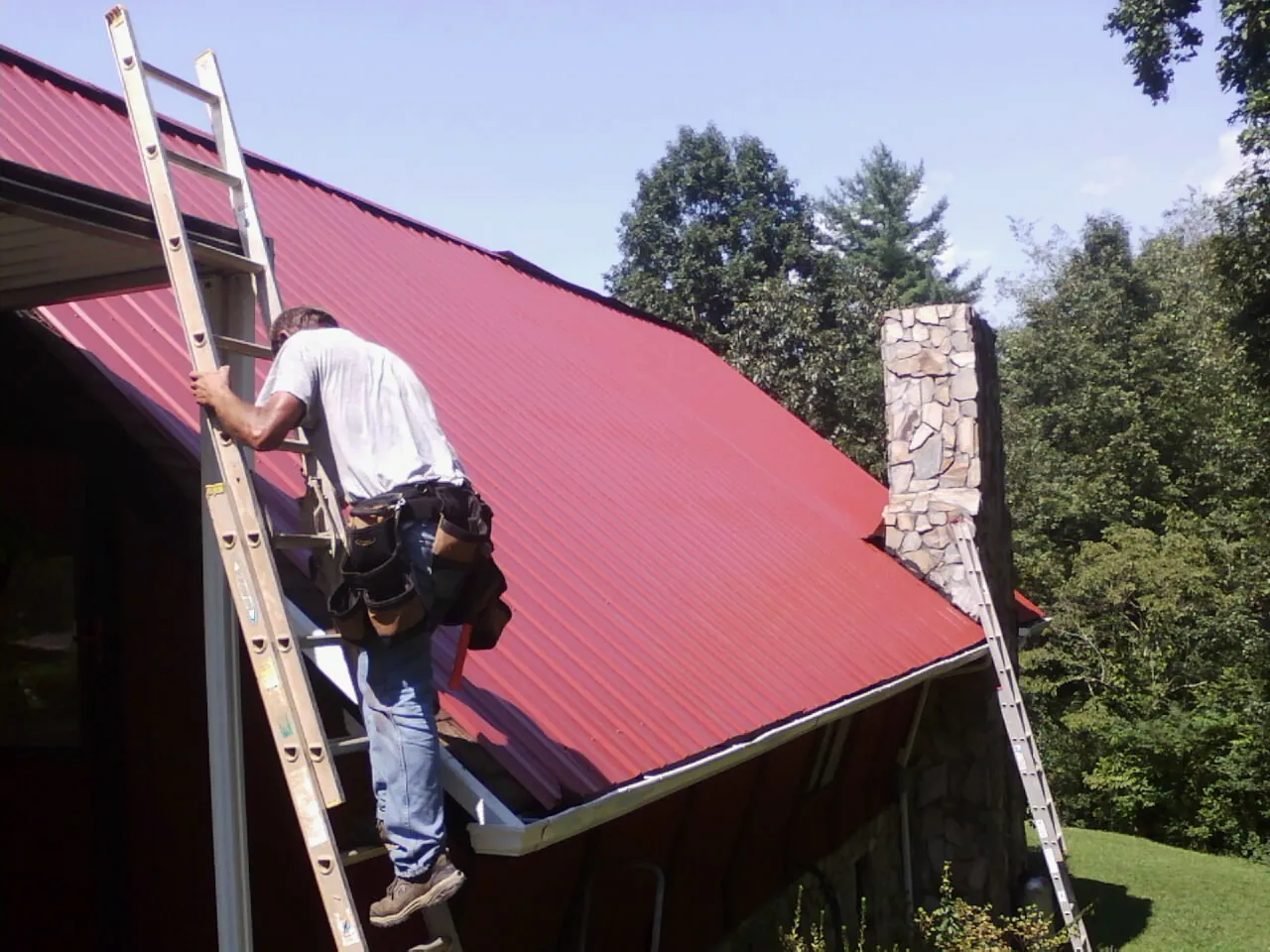 Roofer on a ladder beside a red metal roof and stone chimney — Soffit & Fascia Repair by Roofing Brothers in Pisgah Forest